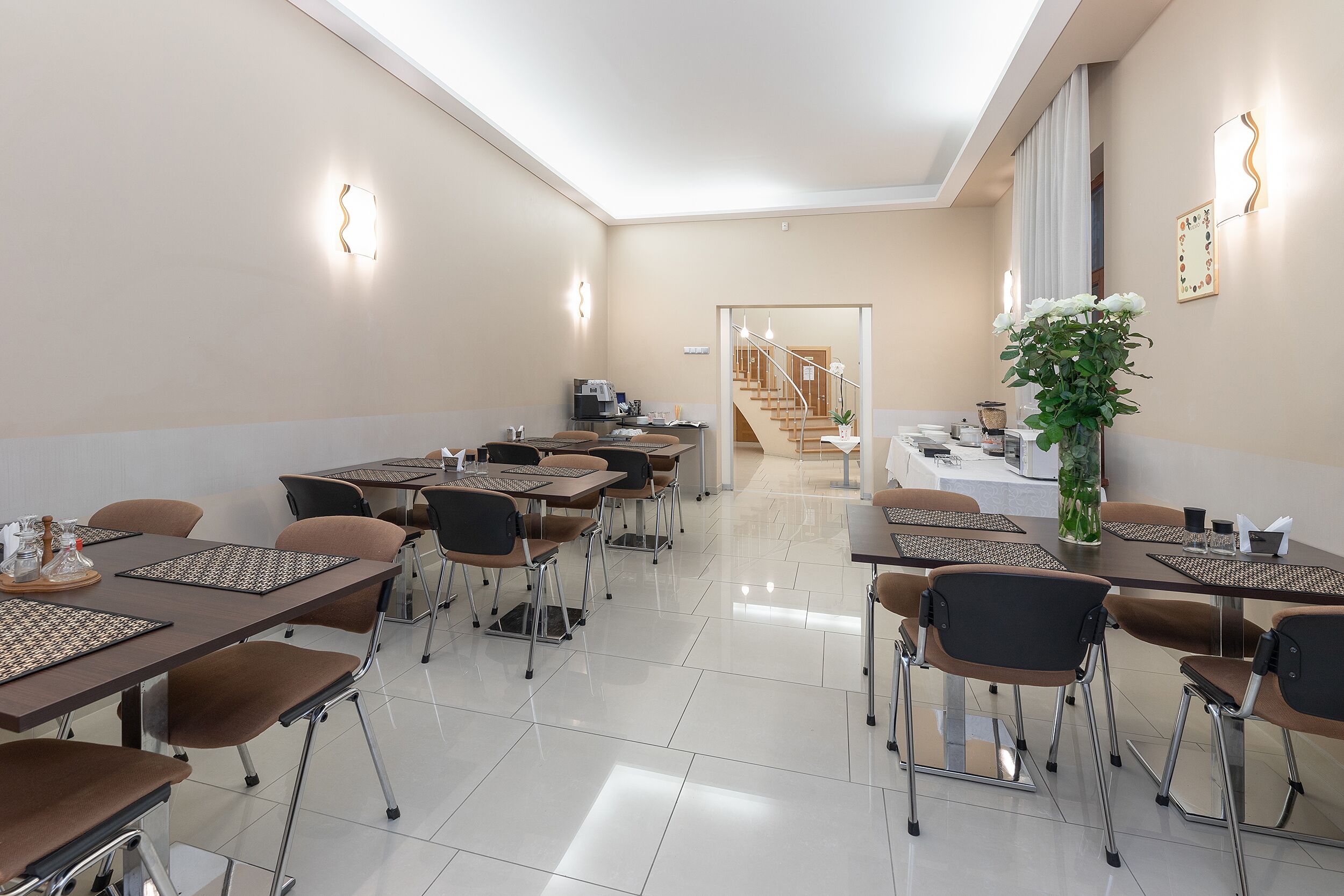 Een restaurantgedeelte in een hotel met meerdere tafels en stoelen, een trap naar een andere verdieping en een grote vaas met bloemen aan de rechterkant.