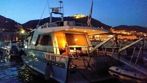 A boat with a covered deck and a cabin area, docked at a marina with buildings in the background. - Akhir Cruise Charter (Salerno)