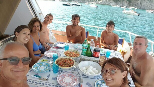 A group of people dining on a boat with a table set for a meal, surrounded by water and boats in the background. - Akhir Cruise Charter (Salerno)