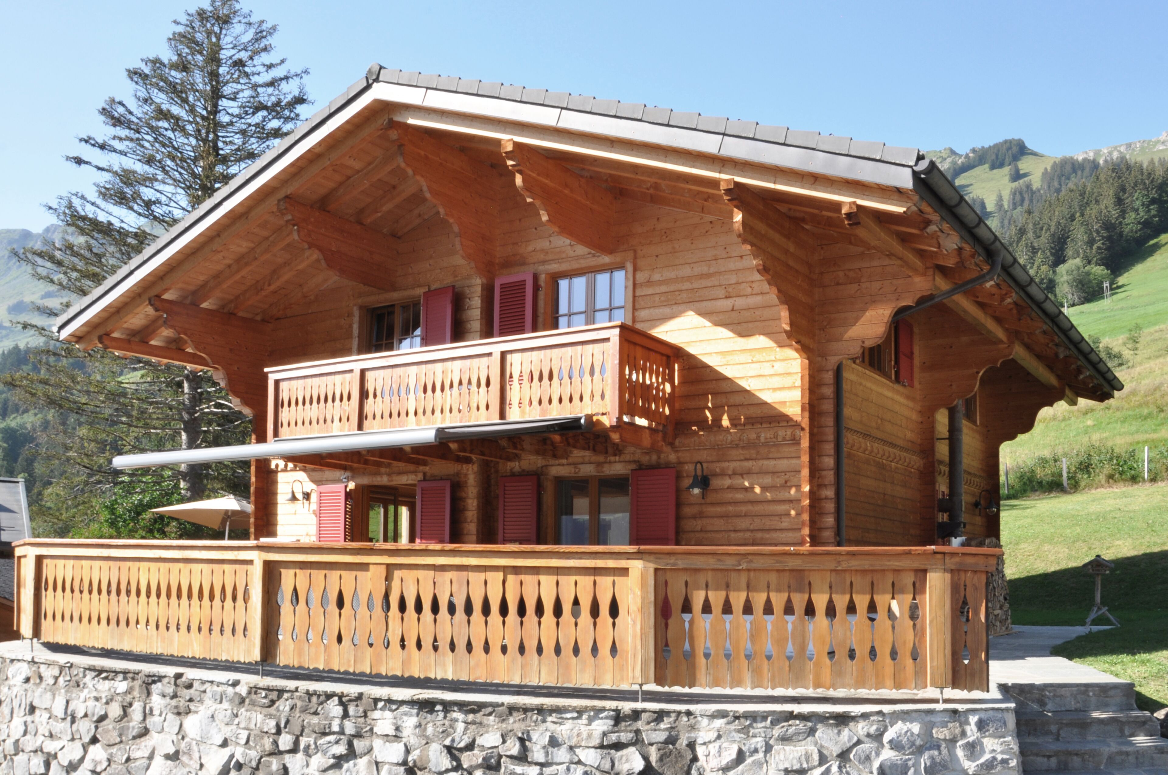 A wooden chalet with a balcony, stone foundation, and a mountainous backdrop.