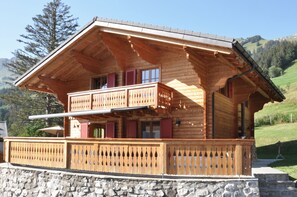 Un chalet en bois doté d’un balcon, avec des fondations en pierre et un paysage montagneux en arrière-plan.