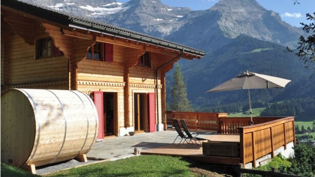 Eine Holzchalet mit einem grossen Fass, einer Terrasse mit Stühlen und Blick auf die Berge.