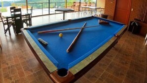 Uma mesa de bilhar com superfície de feltro azul, cercada por cadeiras e mesas, em um salão com piso de azulejos e vista para colinas através de grandes janelas.