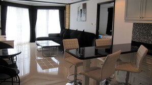 A modern living room with a black sofa, glass dining table, and white chairs. There is a kitchen area with white cabinets and a tiled backsplash.