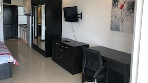 Moderni hotellihuone, jossa on pieni keittiö, seinälle asennettu taulutelevisio ja punakukkia kuvaava taulu.