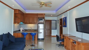 A modern kitchen with wooden cabinets, a microwave, and a refrigerator. There is a blue bar stool, a blue sofa, and a TV mounted on the wall. - Baan Suan Lalana Te Floor 5 Room (Pattaya)