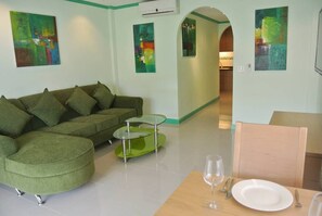 A modern living room with a green sectional sofa, a glass coffee table, and a dining area with a wooden table and chairs.