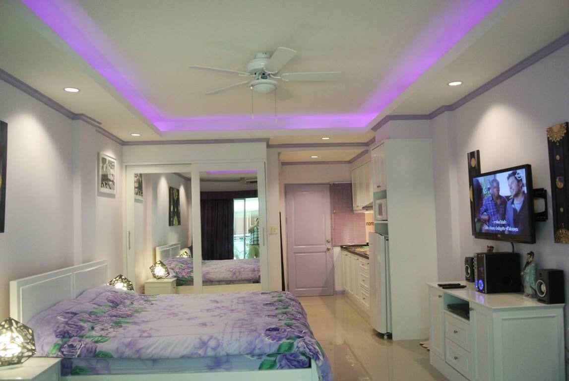 A bedroom with a bed, a ceiling fan, a TV, and a kitchen area.