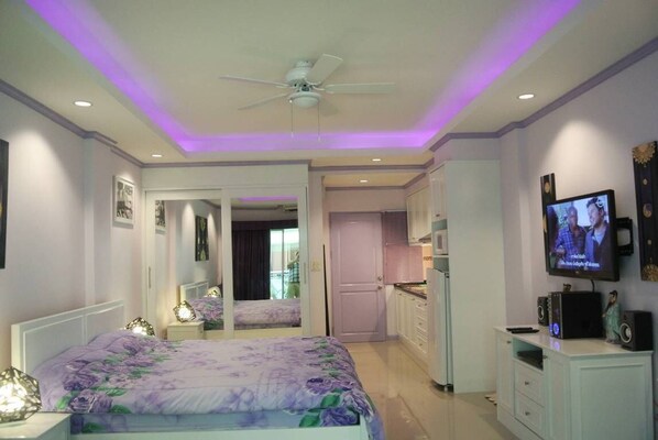 A bedroom with a bed, a ceiling fan, a TV, and a kitchen area.