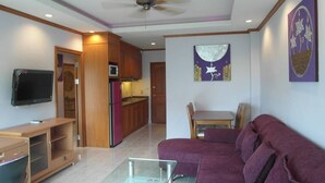 Un soggiorno con un divano viola, un tavolino in vetro, un televisore a schermo piatto, mobili a parete in legno, angolo cottura e una ventola a soffitto.