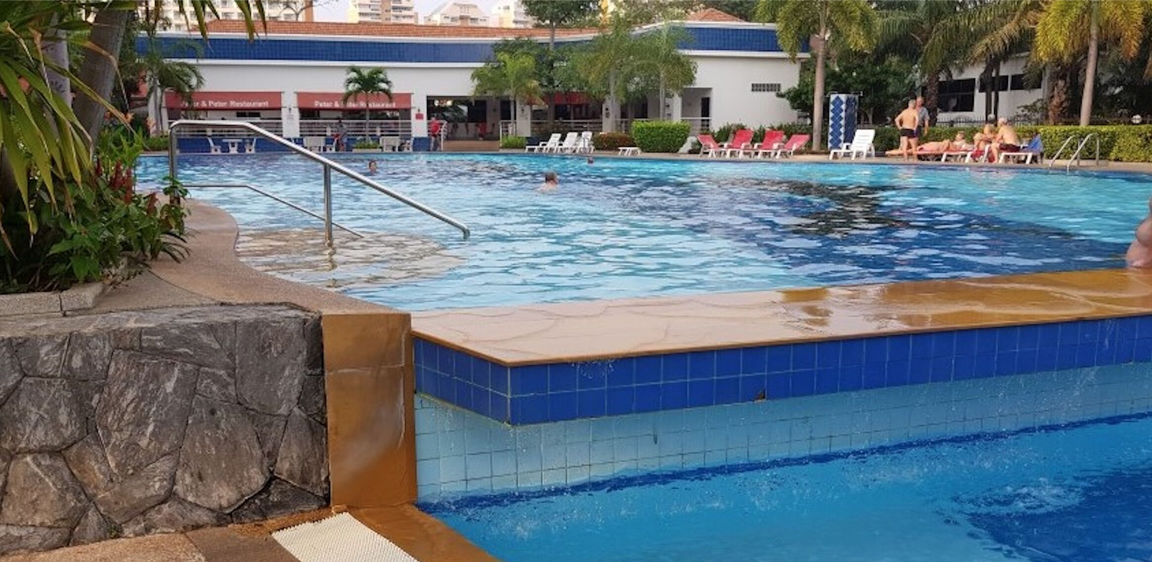 A swimming pool with a stone wall and tiled edge, surrounded by lounge chairs and palm trees.