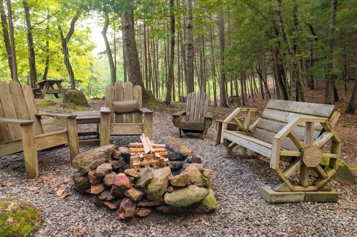 A forest campsite with wooden furniture, a fire pit, and a wooden bench.