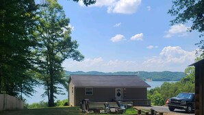A cabin with a view of a lake and hills, outdoor seating, and a vehicle parked nearby.