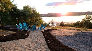 A sandy pathway leading to a seating area with blue chairs and a fire pit, surrounded by trees and a lake view.