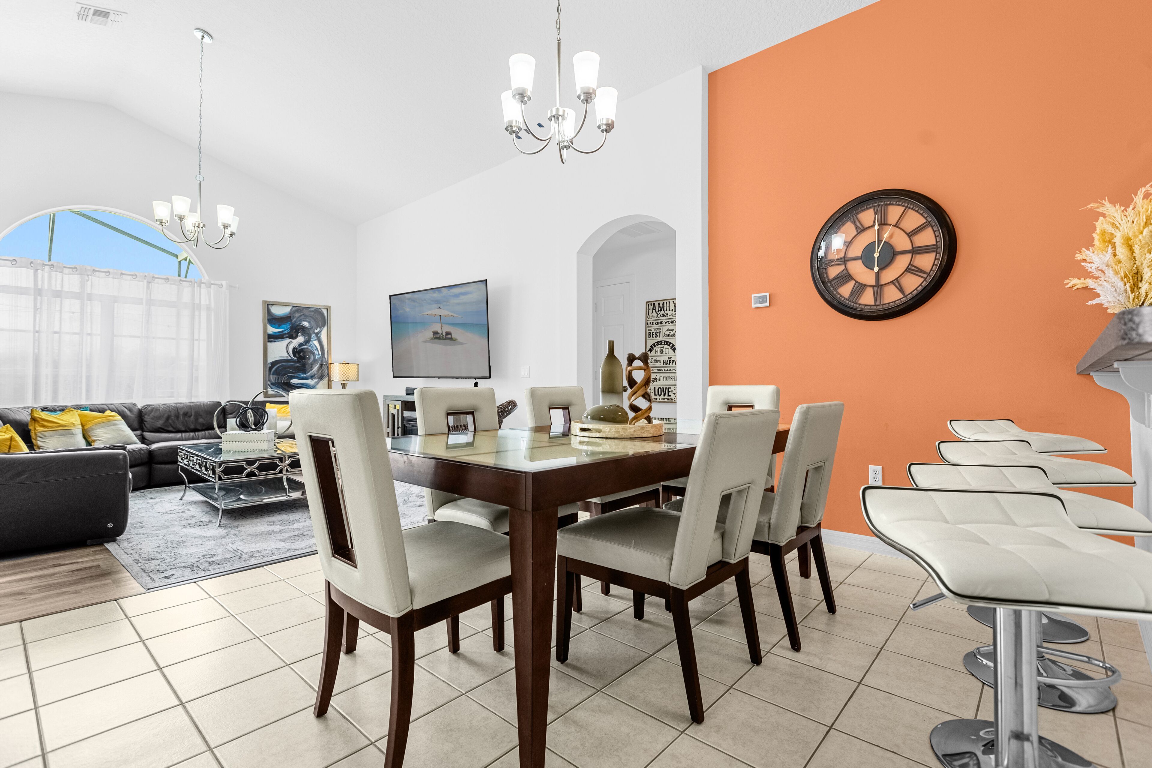A modern living room with a dining area, featuring a large window, a chandelier, and a clock on the wall.