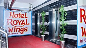 A hotel entrance with a red carpet, a sign for 'Hotel Royal Wings', and two potted plants.