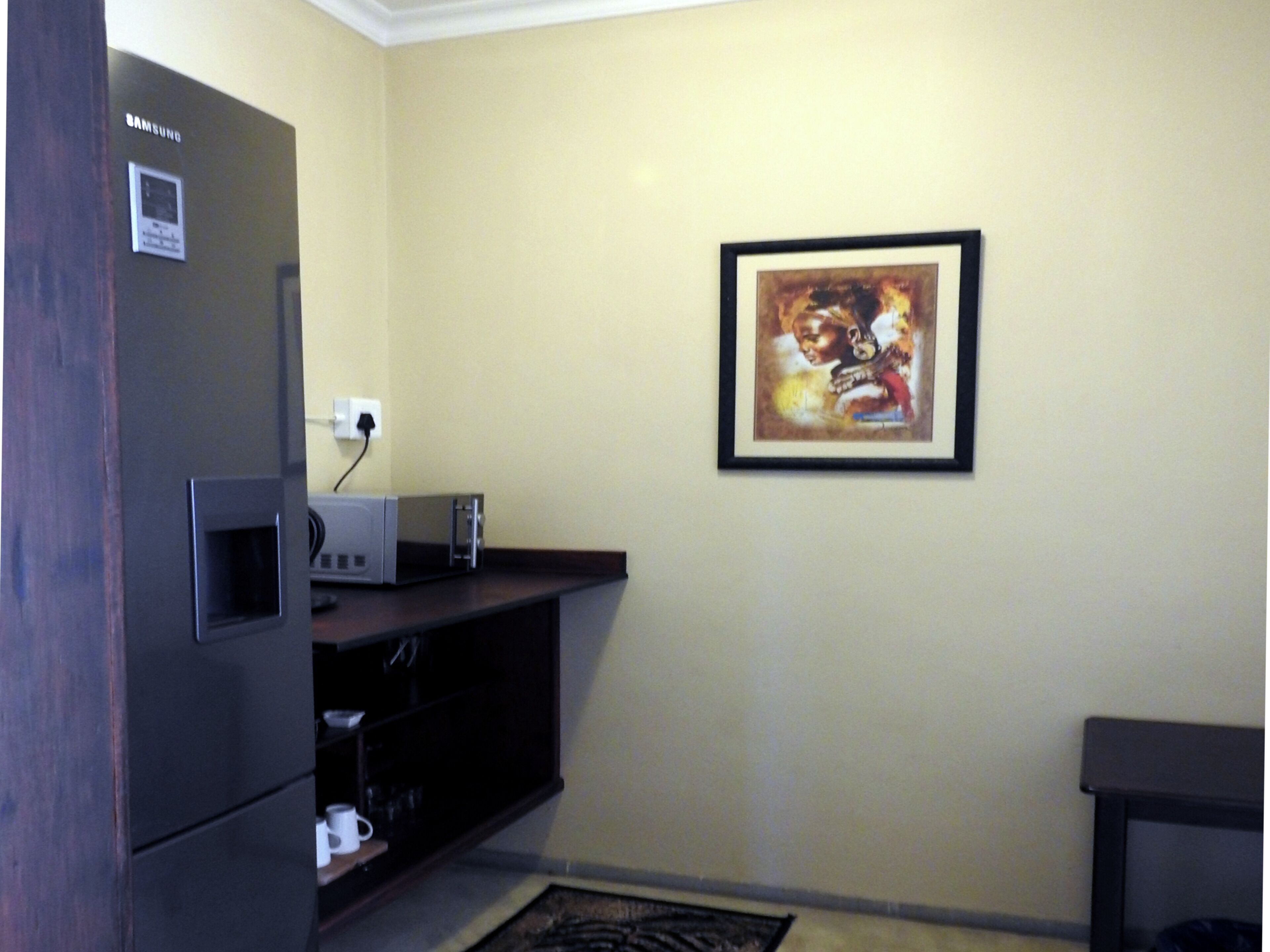 A room with a dark-colored refrigerator, a microwave, and a small cabinet with a few items. There is a framed artwork on the wall and a dark-colored bench or table against the wall.