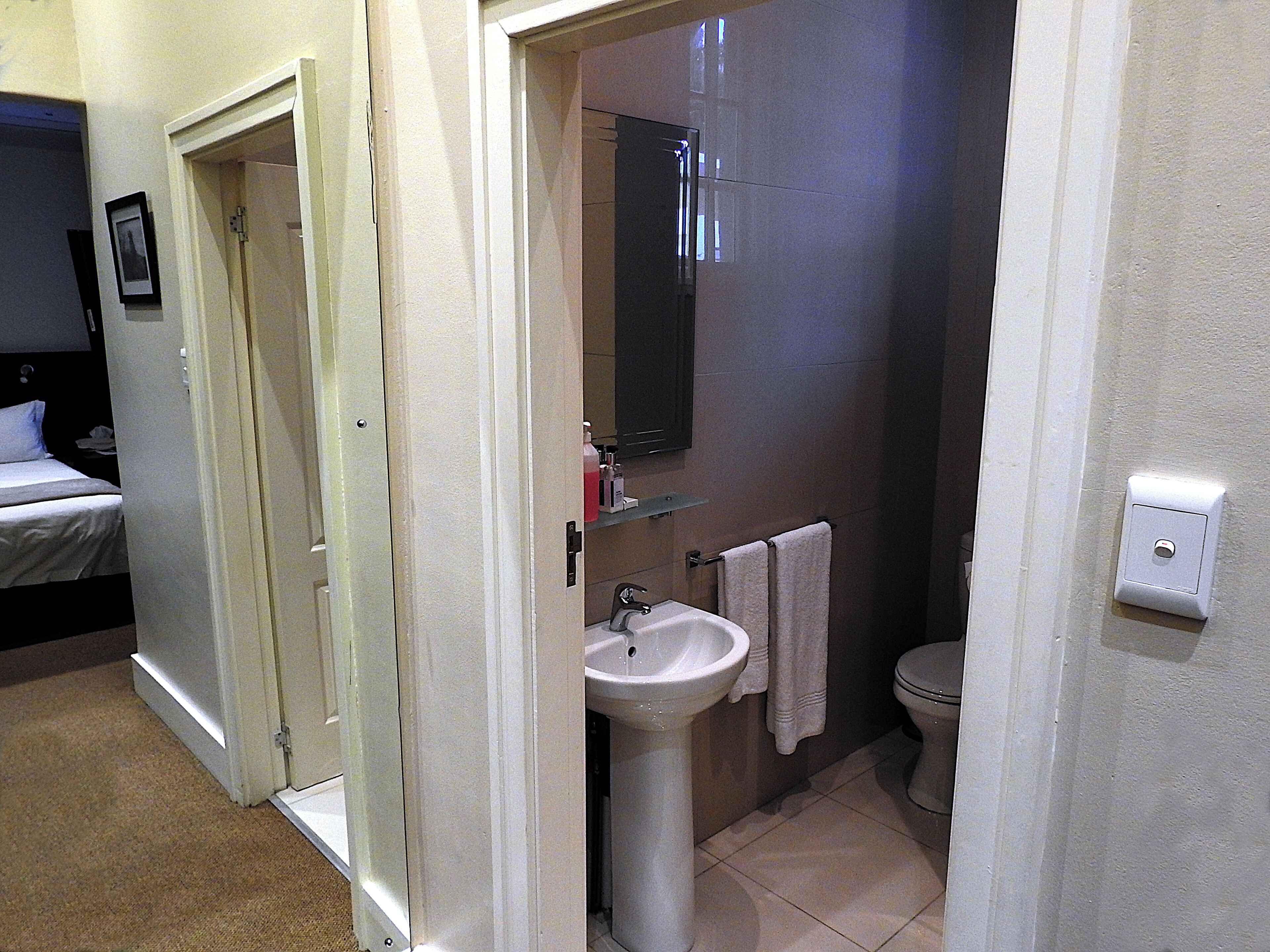 A bathroom with a sink, mirror, and toilet. There is a door leading to a bedroom with a bed.