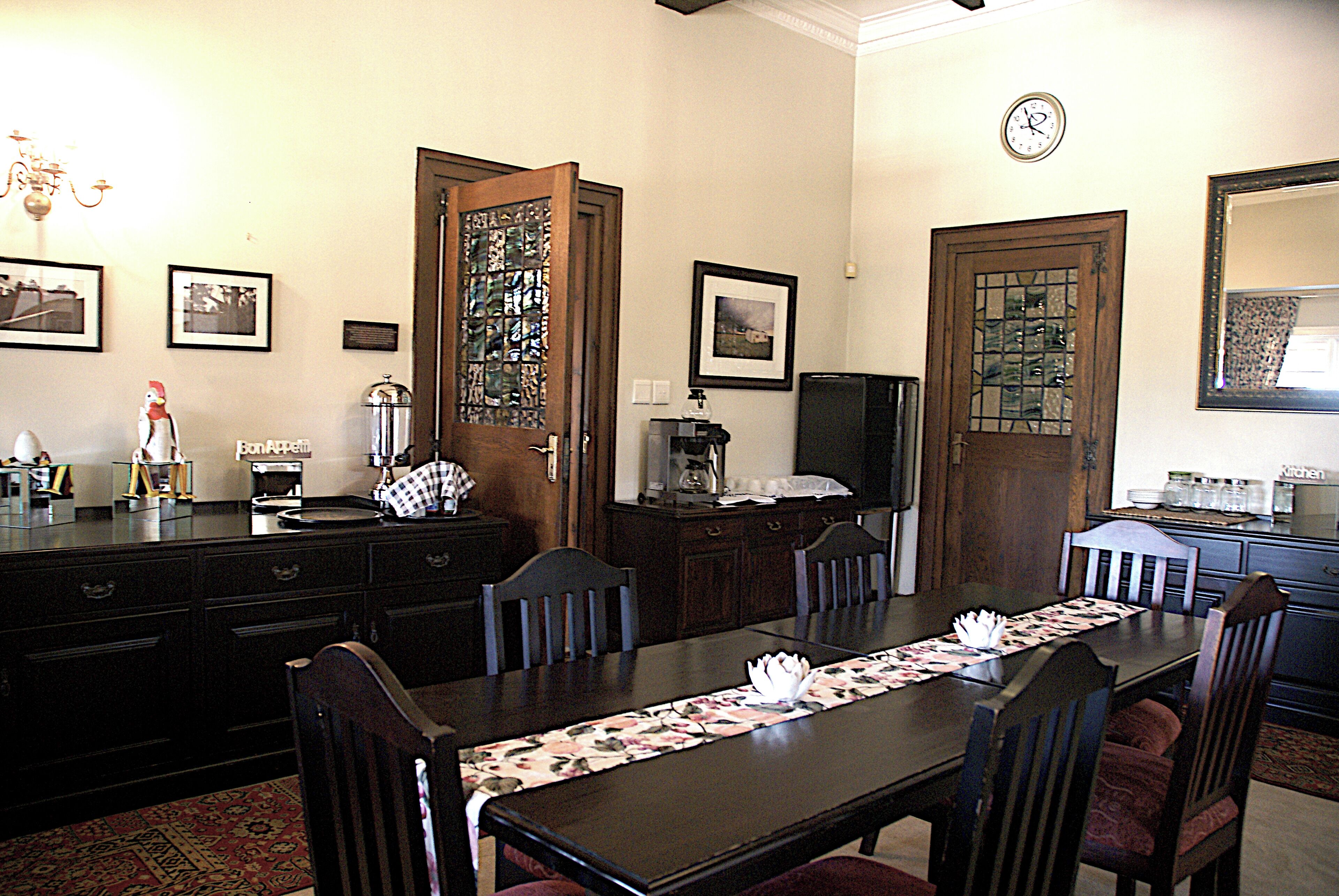A dining area with a long table, chairs, a chandelier, framed pictures, and a clock on the wall.