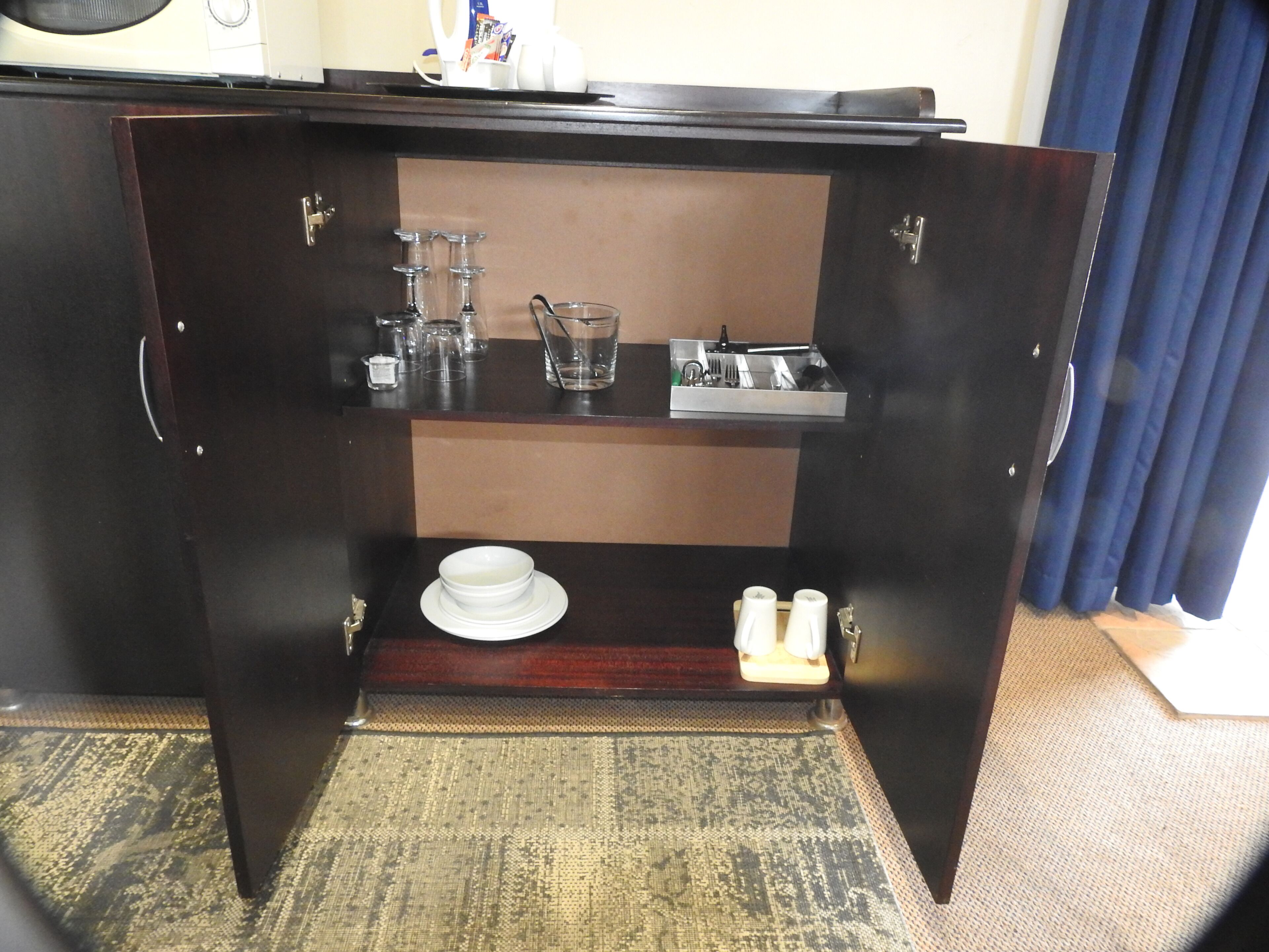 A dark wooden cabinet with a shelf containing a glass, a cup, and a tray with utensils.