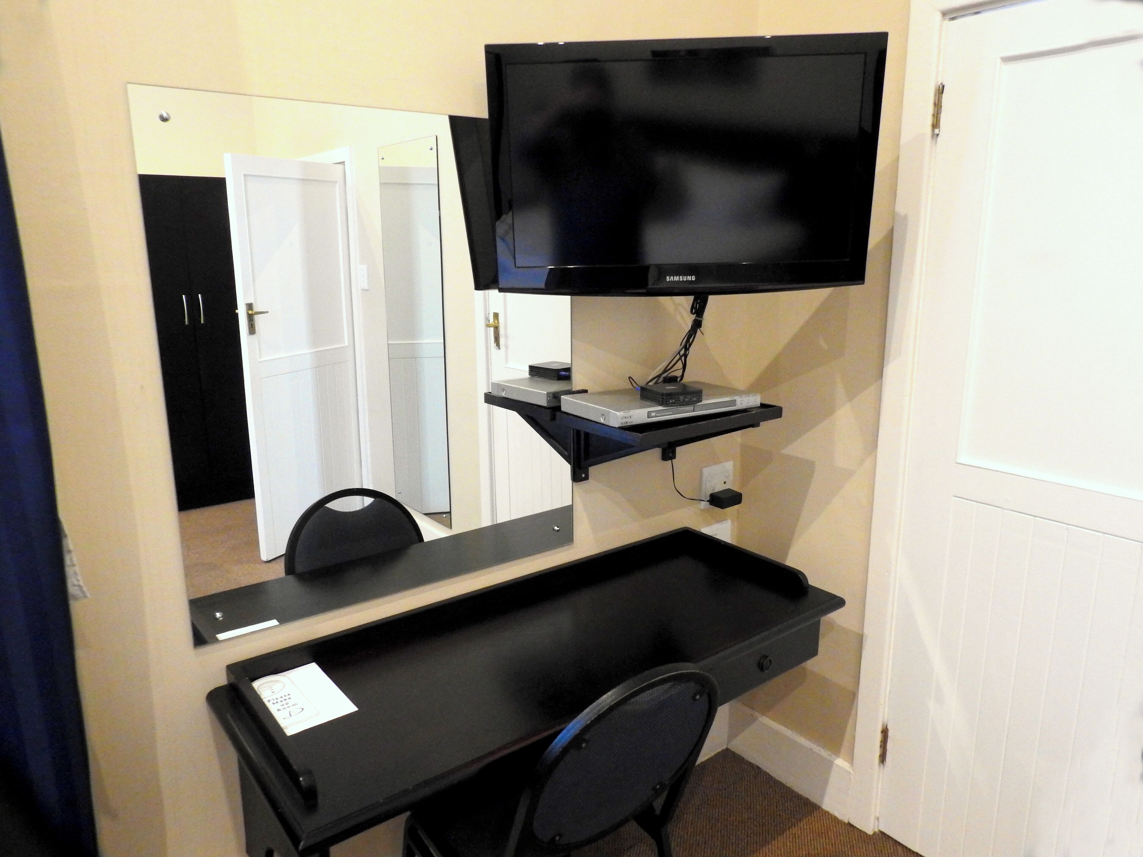 A hotel room with a desk, a flat-screen TV mounted on the wall, a mirror, and a door.