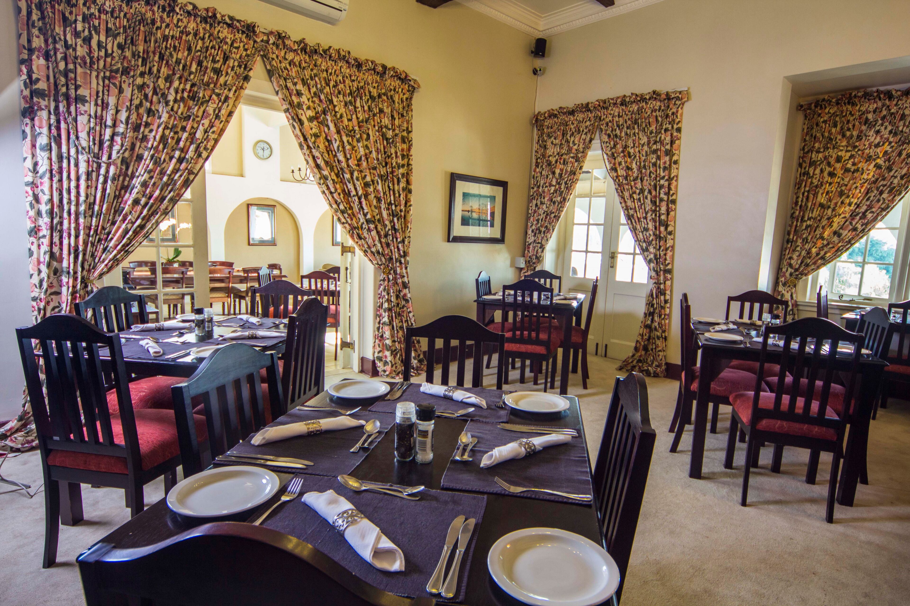 A dining area with tables set for four, each with red upholstered chairs, and floral curtains.