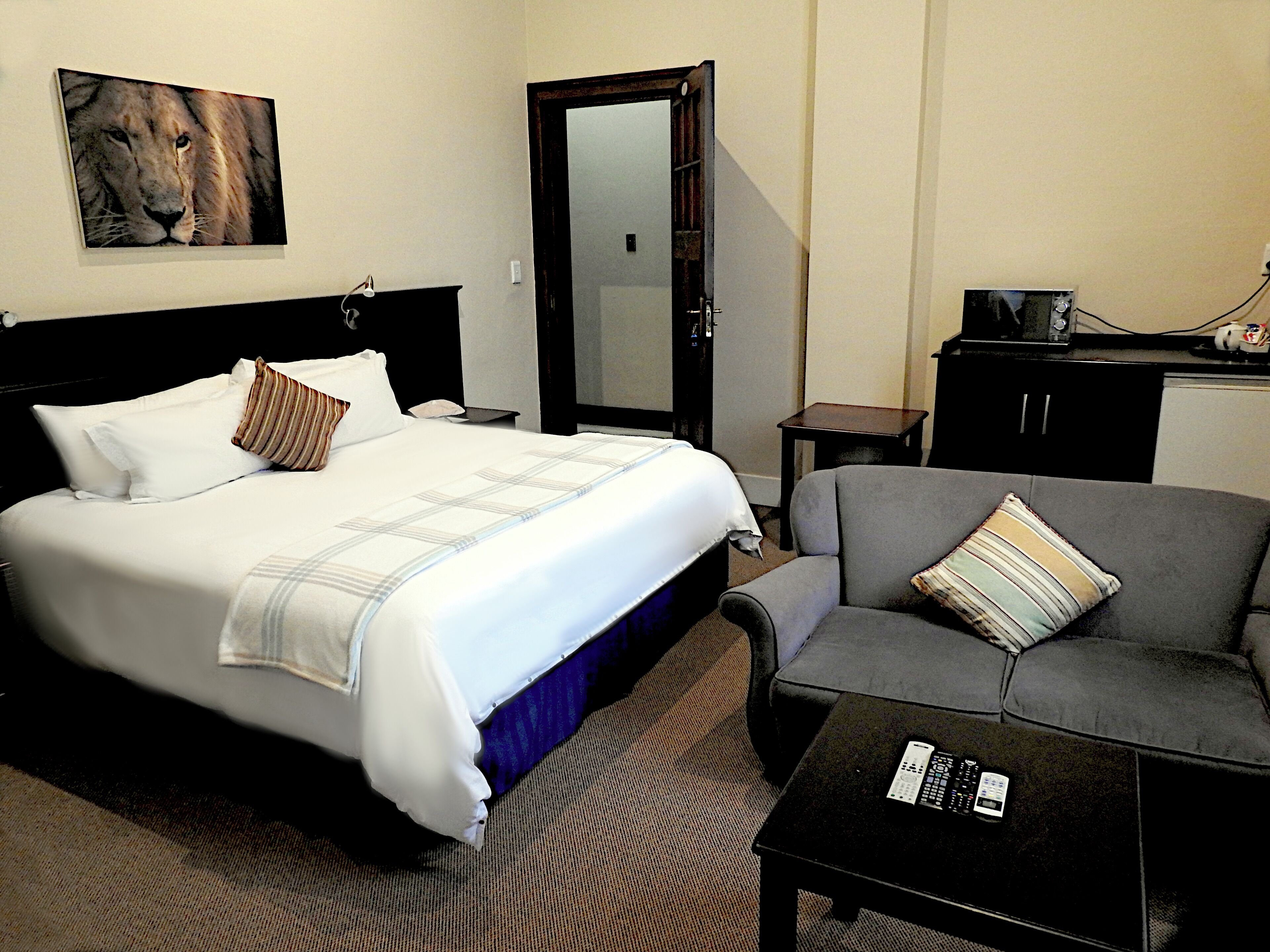 Ein Hotelzimmer mit einem Bett, einem grauen Sofa, einem kleinen Tisch mit einer Fernbedienung, einem Gemälde eines Löwen und einer Tür zu einem anderen Raum.