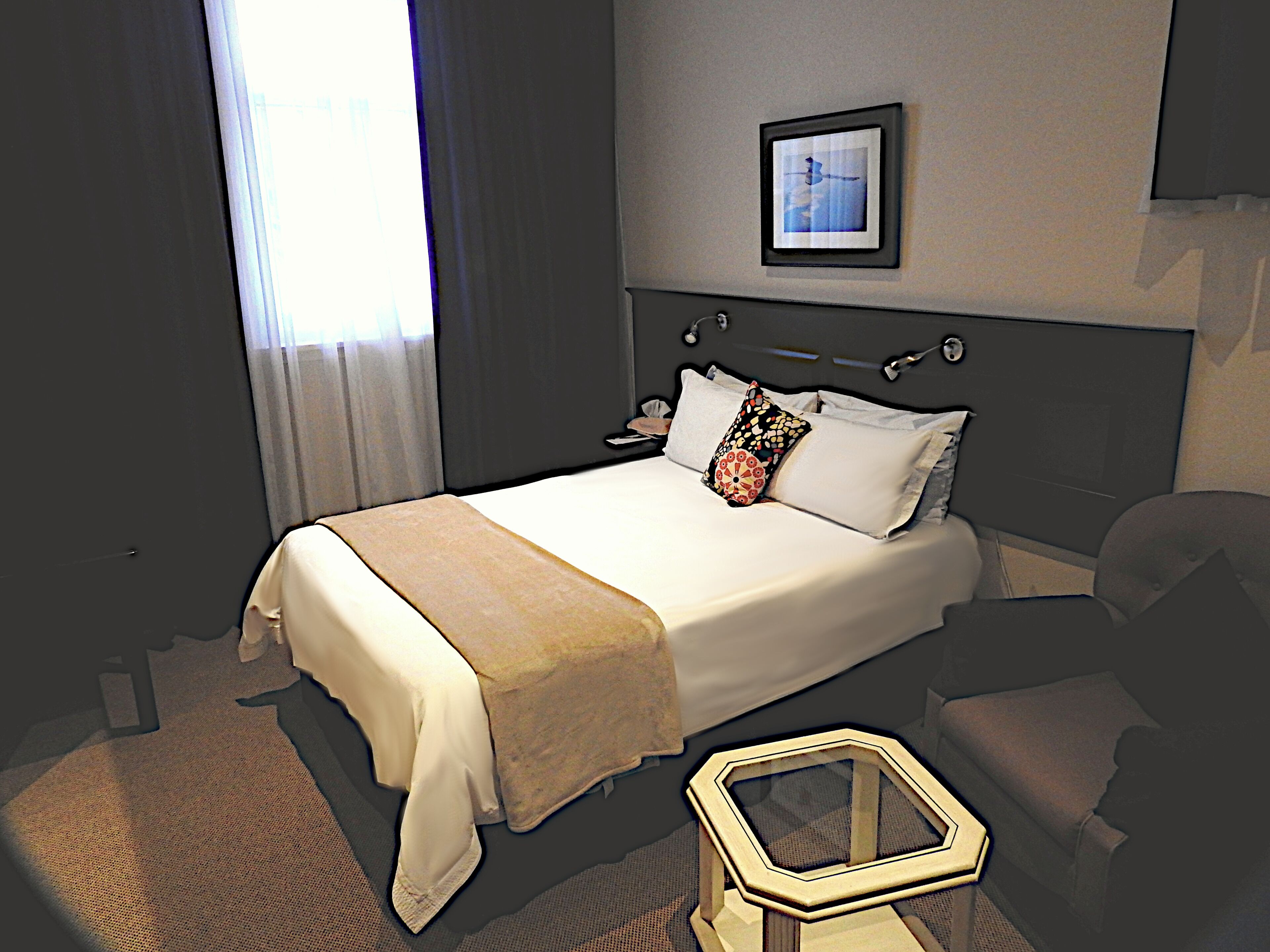 Quarto de hotel com cama, mesa de cabeceira, uma cadeira e uma pequena mesa com tampo hexagonal.