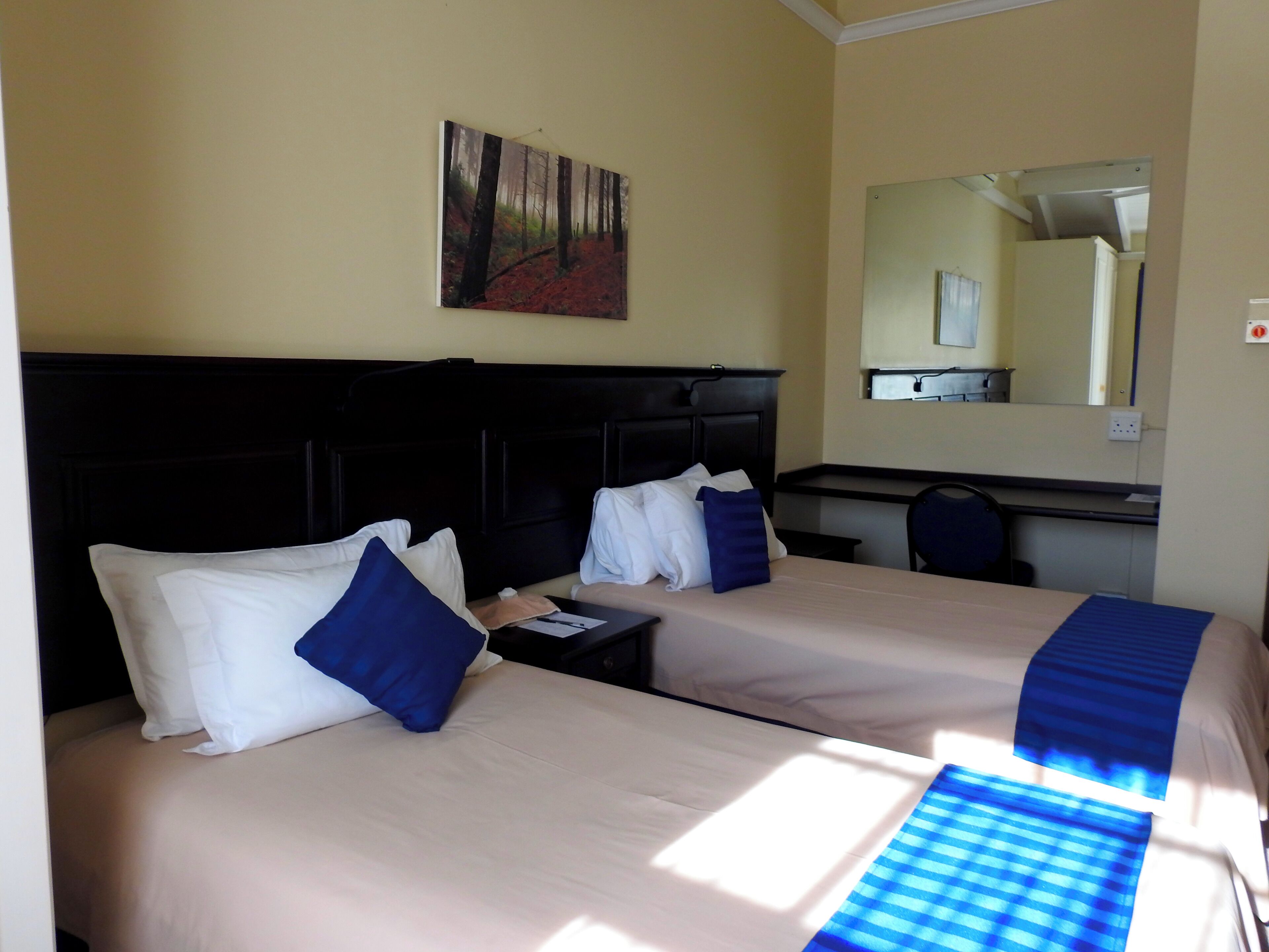 A hotel room with two beds, a desk, a chair, a painting on the wall, and a mirror above the desk.
