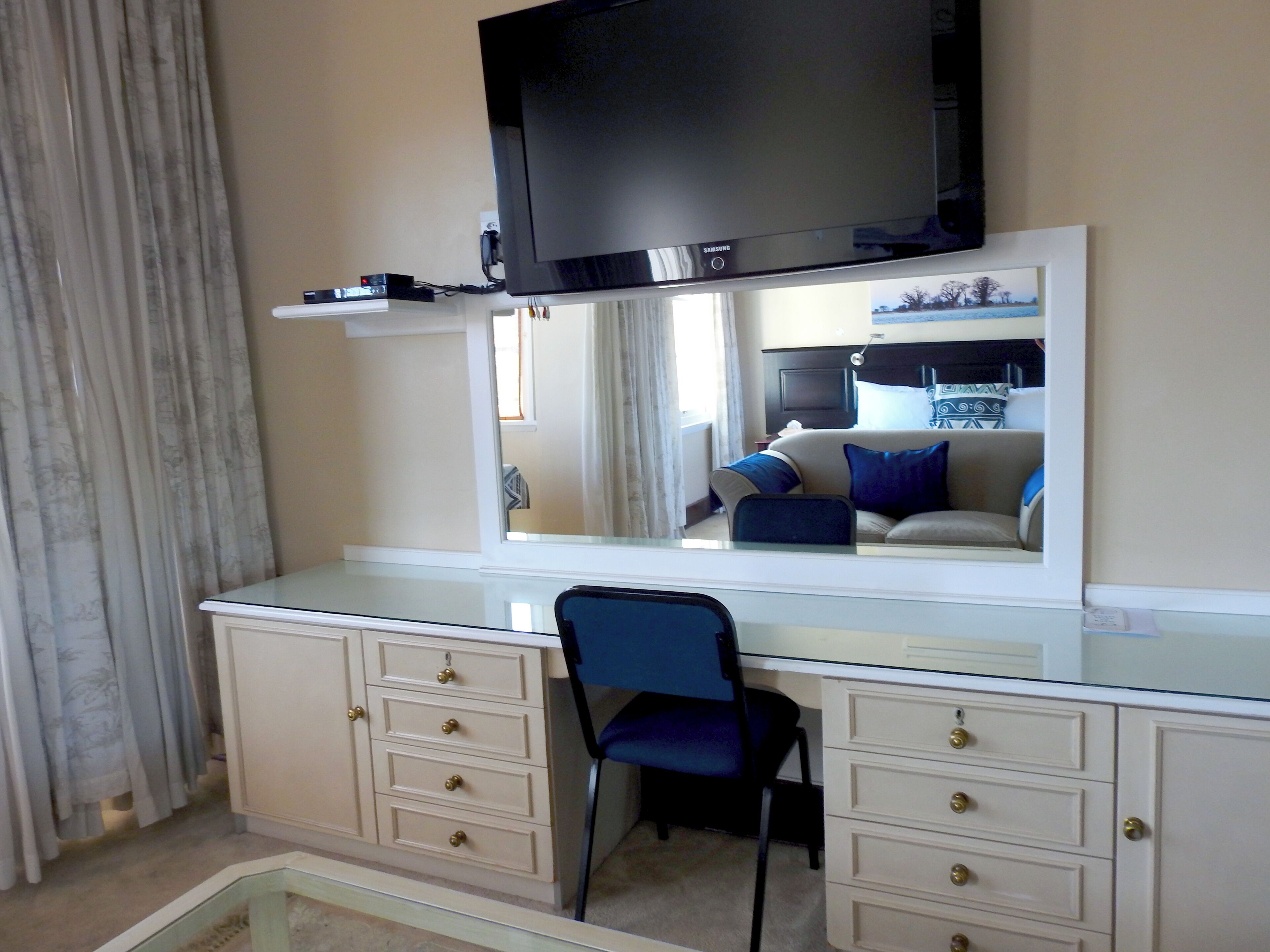 A hotel room with a large flat-screen TV mounted above a glass desk, a chair, and a dresser with drawers.