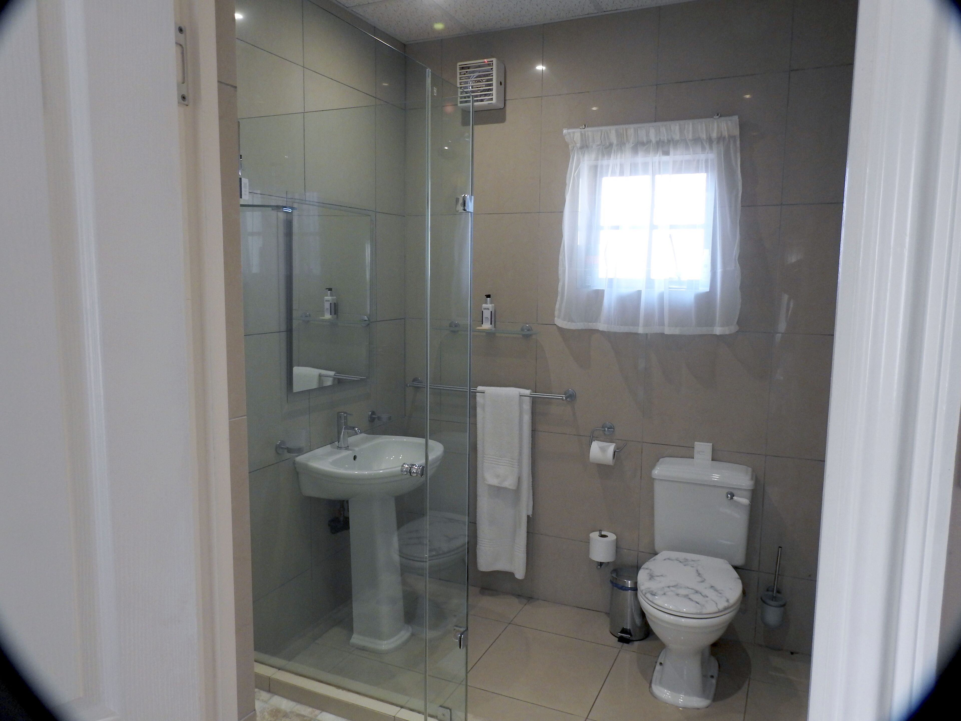 A modern bathroom with a glass shower enclosure, a pedestal sink, a toilet, and a window with sheer curtains.
