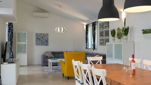 Uma sala de estar moderna com um sofá, uma poltrona amarela, uma mesa de jantar com cadeiras e uma área de cozinha com armários e plantas.