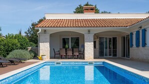Uma casa de dois andares com piscina, pilares de pedra e telhado em telhas.