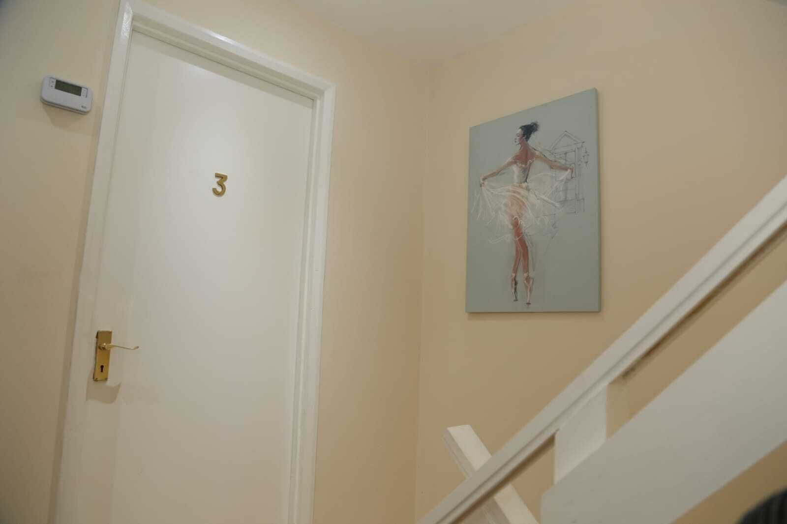 A hallway with a white door marked with the number 3, a thermostat on the wall, and a painting of a dancer.
