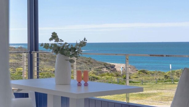 Un balcon donnant sur l’océan, un vase blanc orné de verdure et deux objets roses.