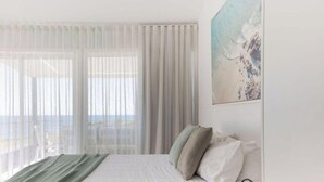 Une chambre à coucher avec un lit, des rideaux blancs, un tableau et une vue sur la mer.