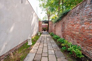 A narrow pathway flanked by a brick wall and a white wall, leading to a covered entrance.