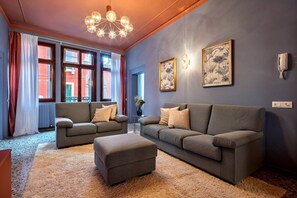 A living room with two sofas, a coffee table, and a chandelier. There are windows with curtains and a wall-mounted phone.