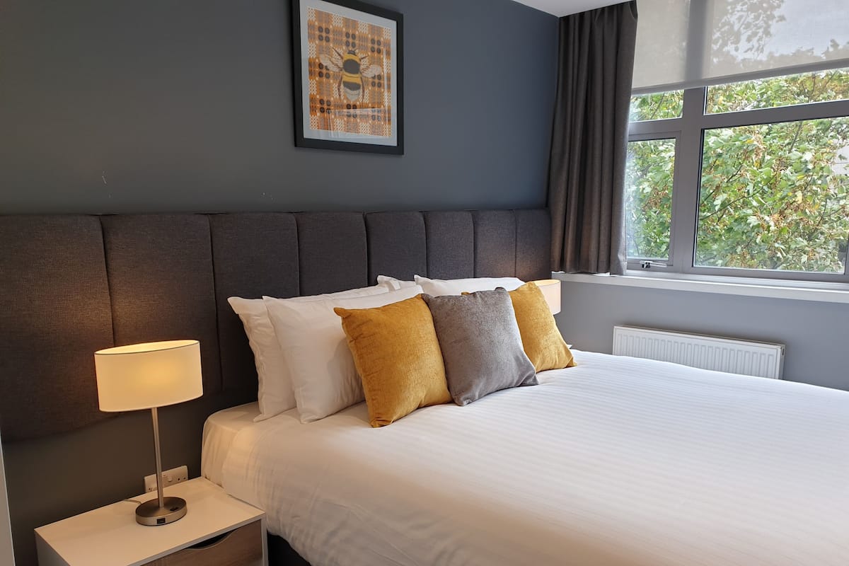 A neatly made bed with white and mustard pillows, a bedside lamp, and a framed artwork on the wall.