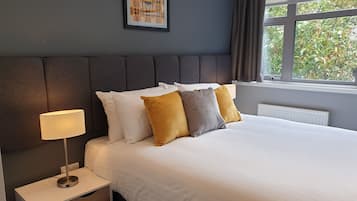A neatly made bed with white and mustard pillows, a bedside lamp, and a framed artwork on the wall.