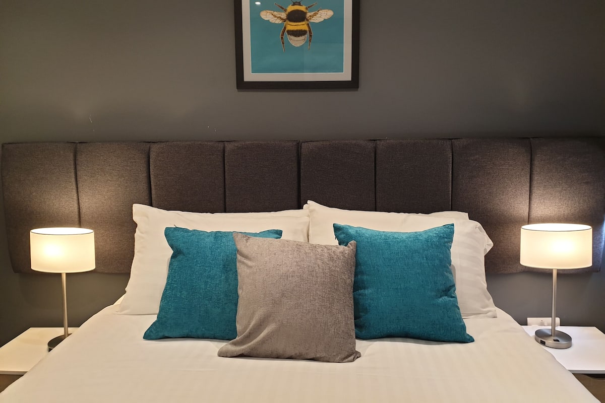 A neatly made bed with white and teal pillows, a gray accent pillow, and a framed artwork of a bee on the wall above.