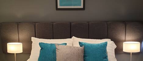 A neatly made bed with white and teal pillows, a gray accent pillow, and a framed artwork of a bee on the wall above.