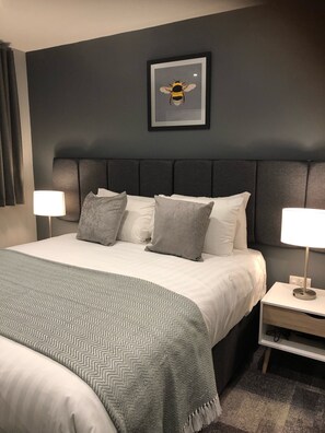 A modern hotel room with a large bed, grey and white bedding, a grey headboard, two bedside lamps, and a framed bee artwork on the wall. - The Spires Serviced Apartments Cardiff (Cardiff)