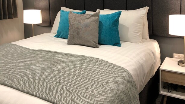 Habitación de hotel con una cama grande, almohadas grises y azules, una obra de arte con temática de abeja en la pared y lámparas de noche.
