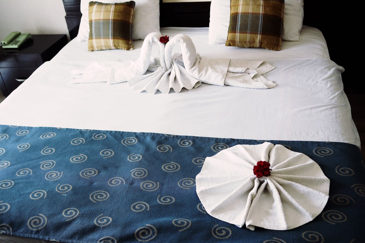 A neatly made bed with a towel swan and a towel flower on a blue patterned bedspread.