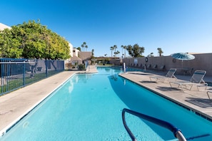 Piscina azul e límpida com cadeiras de descanso e guarda-sóis, cercada por um muro azul e palmeiras.