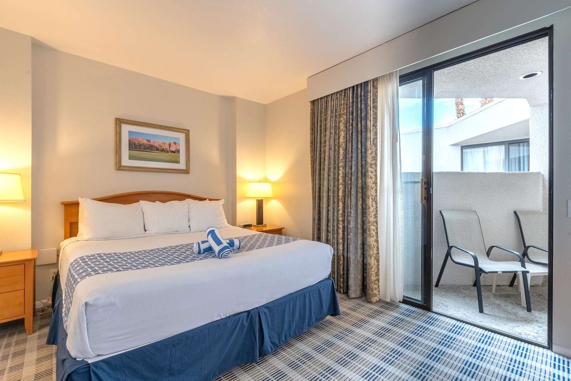 A hotel room with a bed, two bedside lamps, a framed picture on the wall, and a view of a balcony with a chair.