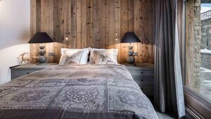 A bedroom with a wooden ceiling, a large bed with patterned bedding, two bedside lamps, and a view of a snowy landscape through a window.
