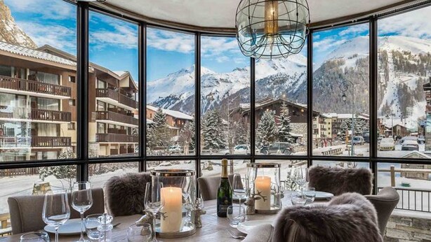 A dining area with a table set for two, large windows offering a view of snow-covered mountains and a town below.