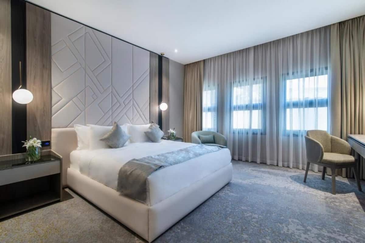 Ein modernes Hotelzimmer mit einem großen Bett, einem Schreibtisch, einem Sessel und Blick auf die Stadt durch große Fenster.